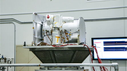 A high-tech device is displayed on a platform in a lab, with a monitor showing data in the background.