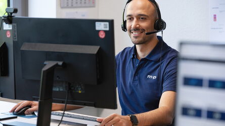 A man wearing a headset smiles while providing support at his computer workstation.