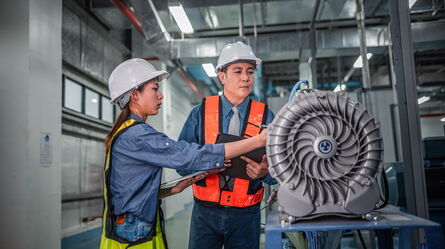 Two engineers in hard hats and vests discuss machinery plans in an industrial setting.