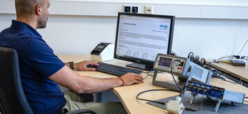 A man in a blue shirt is reviewing a calibration certificate on a computer screen at a desk with electronic equipment.