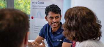 A consultant engages with colleagues in a meeting, gesturing while discussing ideas in a bright office setting.