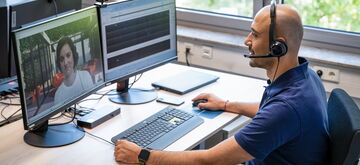 A man in a headset provides customer support via video call on dual monitors in a bright office setting.