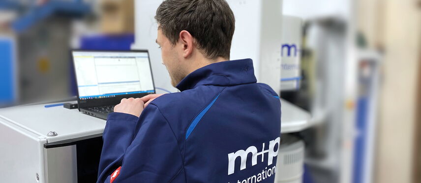 A technician in a blue MHP International jacket works on a laptop at a testing station.