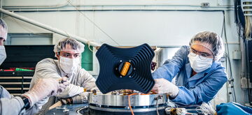 Technicians in protective gear work on a complex machinery setup in a laboratory environment.