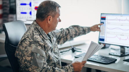A person in military uniform reviews data trends on a computer screen, pointing and holding documents in a monitoring room.