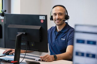 A smiling customer support representative wearing a headset sits at a desk with dual monitors.