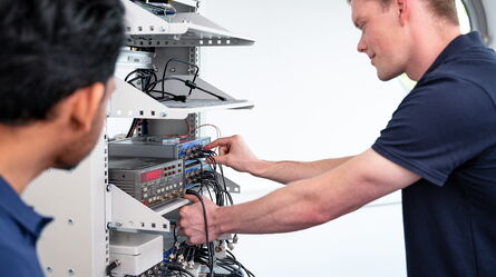 Two technicians in navy shirts work on wiring and installation inside a metal server rack.