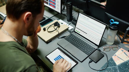 A man monitors data on a laptop and tablet at a desk, focusing on maintaining safety and high-uptime operations.