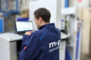 A technician in an "m+p international" jacket works on a laptop at a test stand in an industrial setting.