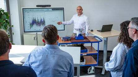 A man in a white shirt conducts a training session, explaining data on a screen to an attentive group.