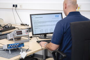 A man in a blue shirt reviews a calibration certificate on a computer screen in a technical office setting.