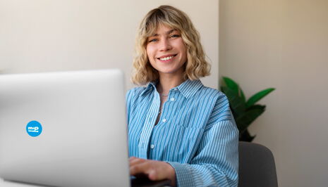 A woman in a striped shirt smiles while working on a laptop, with a plant in the background.
