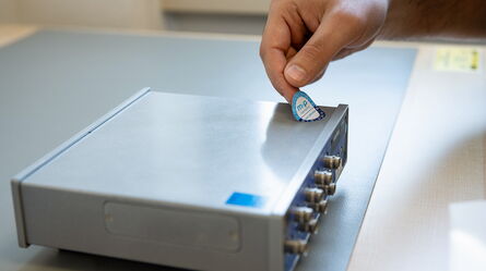 A hand applies a calibration sticker to a metal device, ensuring ISO IEC 17025 compliance and service support.