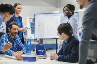 A team collaborates around a table with electronic equipment and a screen displaying technical diagrams during a training session.