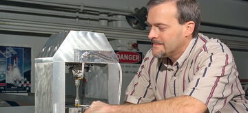 A man in a striped shirt adjusts a scientific instrument inside a metallic enclosure in a laboratory setting.