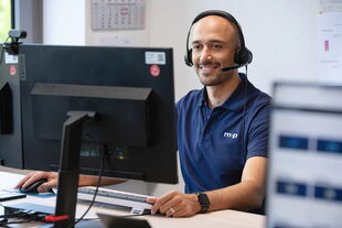 A man in a headset smiles while providing tech support at a computer, embodying professional assistance.
