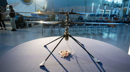 A small helicopter model with long blades is displayed on a table in an exhibition hall.
