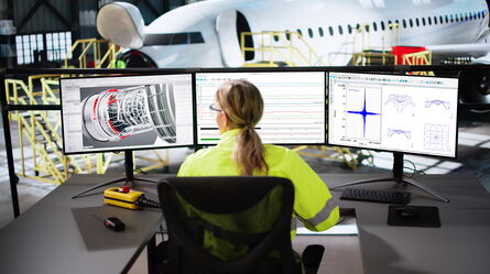 A technician in a high-visibility jacket monitors aircraft data on three screens in a hangar, enhancing workflow automation.
