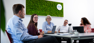 A group of five people engage in a meeting around a table, with a green wall art piece displaying the logo "m+p international."