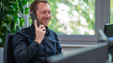 A smiling man in a black shirt engages in a customer support call in a bright office setting.