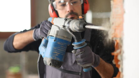 A man wearing protective gear uses a power drill to chip away at a brick wall, demonstrating testing durability.