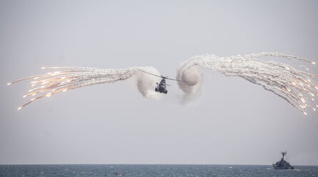 A military helicopter deploys flares over the ocean, demonstrating defense standards amidst a cloudy sky.