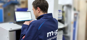 A technician in an MHP jacket works on a laptop at a testing station in an industrial setting.