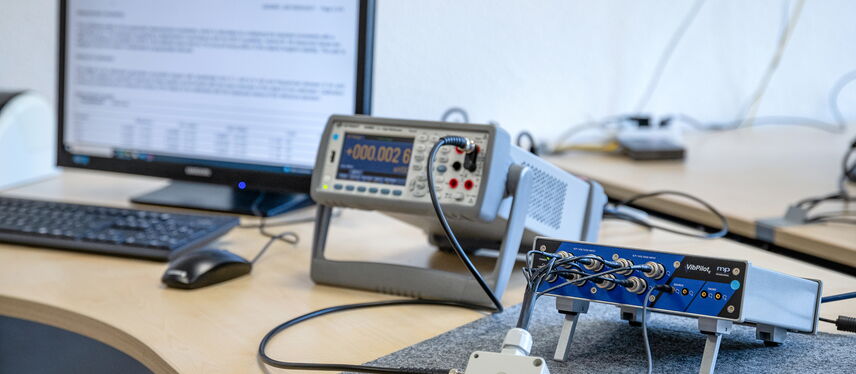 A calibration setup with measuring instruments and a computer displaying a certificate is arranged on a wooden desk.