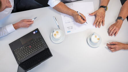 Three people collaborate at a desk with a laptop, documents, and cappuccinos, discussing a work-related quote.