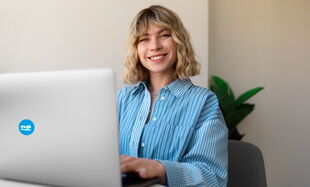 A woman in a striped shirt smiles while working on a laptop, with a plant in the background.