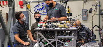 Technicians in masks work collaboratively on a complex machinery setup in a high-tech laboratory environment.