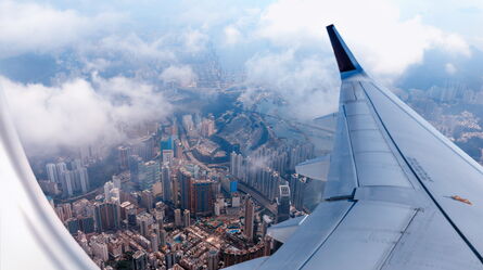 An airplane wing soars above a bustling cityscape, dotted with skyscrapers and clouds, illustrating aviation dynamics.