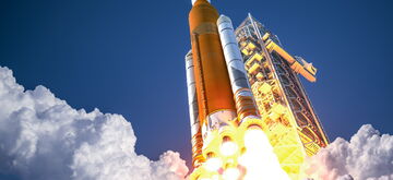 A spacecraft launches into a clear sky, leaving behind a trail of smoke and fire during its qualification phase.