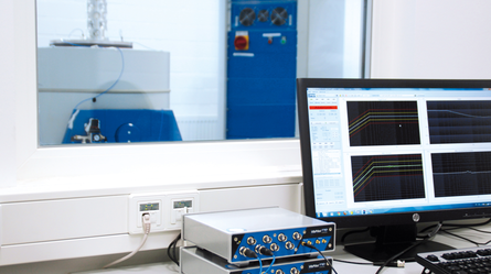 A computer screen displays data graphs in a lab, with equipment visible through a window in the background.