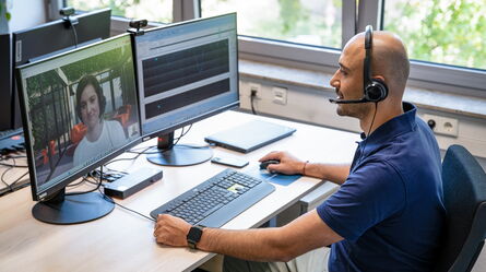 A customer support representative wearing a headset assists a client via video call at a dual-monitor workstation.