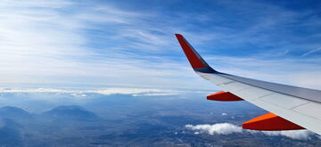 An airplane wing soars over a vast landscape, with mountains and clouds under a clear blue sky.