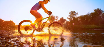 A person rides an electric bike through a shallow river at sunset, creating a dynamic splash.