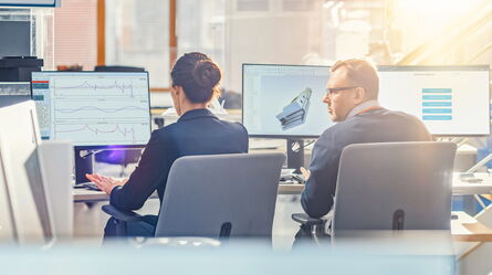 Two professionals discuss data trends and 3D models on multiple monitors, enhancing workflow automation efficiency.