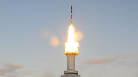 A rocket launches from a snow-covered platform, emitting bright flames and smoke into the clear blue sky.