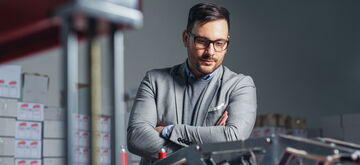 A man in a gray suit observes machinery in an industrial warehouse, reflecting a focused work environment.