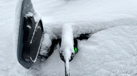 A snow-covered electric vehicle charges, with a green light indicating active power flow amidst wintry conditions.