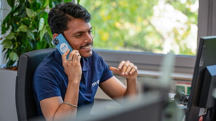 A service engineer in a blue polo shirt talks on a smartphone while sitting at his desk, surrounded by office plants.
